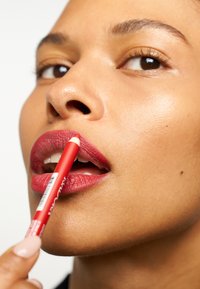 Red lip pencil held near lips, matte finish on pursed lips, smooth skin texture, natural light highlighting contours, minimal background.