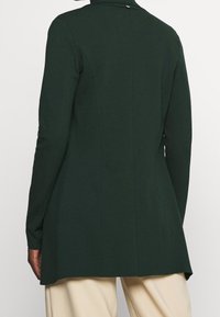 Dark green fitted blazer with a smooth texture, slightly elongated back, and subtle stitching details, designed for a tailored look.