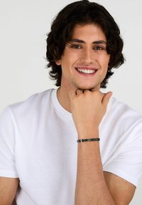 Bracciale in metallo nero con anelli a catena intrecciati, indossato su un polso. Il modello ha i capelli mossi e indossa una t-shirt bianca a girocollo.