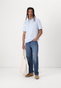 Lysblå strikket polo med vandrette striber, parret med blå denimjeans og beige sko, mens der holdes en beige canvas tote.