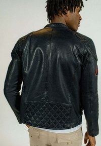 Veste de moto en cuir noir avec texture matelassée dans le dos, épaules rembourrées et panneaux latéraux. Présente un écusson sur le haut de la manche.