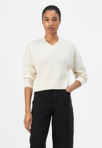 Pull en tricot couleur crème avec un col en V et des poignets côtelés ; présente un petit patch en forme de cœur sur la poitrine gauche ; associé à un jean noir.