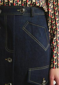 Jupe en denim foncé avec des coutures jaunes contrastantes et de grandes poches avant. Des boutons en argent ornent la taille, ajoutant un détail fonctionnel.