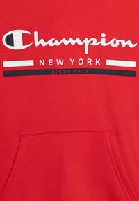 Tissu rouge avec le logo Champion en blanc et noir, texte "New York" et slogan "Depuis 1919" au-dessus d'une poche avant.