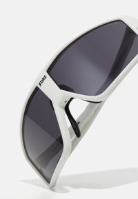 Weiße übergroße Sonnenbrille mit einem geometrischen Gestell, dunkelgrauen Gläsern, gummierten schwarzen Akzenten an den Bügeln und Branding an der Seite.