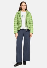 Zielona kurtka puchowa z kapturem, zapinana na zamek błyskawiczny i zatrzaski, zestawiona z białym swetrem i granatowymi spodniami o szerokich nogawkach w paski.