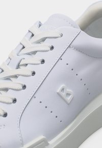 Baskets en cuir blanc avec lacets blancs, détails perforés sur les côtés, et un logo "B" en relief près de la semelle sur le côté extérieur.