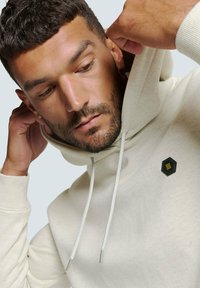 Lichtbeige hoodie met een gestructureerde stof, verstelbare koordjes en een zwart hexagonaal logo-op-de- borst met een geel logo.