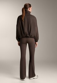 Brown puffer jacket with a high collar, elastic waist, and dropped shoulders paired with matching flared leggings and white sneakers.