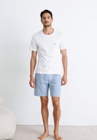 T-shirt blanc à manches courtes avec un petit logo noir, associé à un short à motif bleu clair. Le modèle se tient pieds nus contre un fond uni.