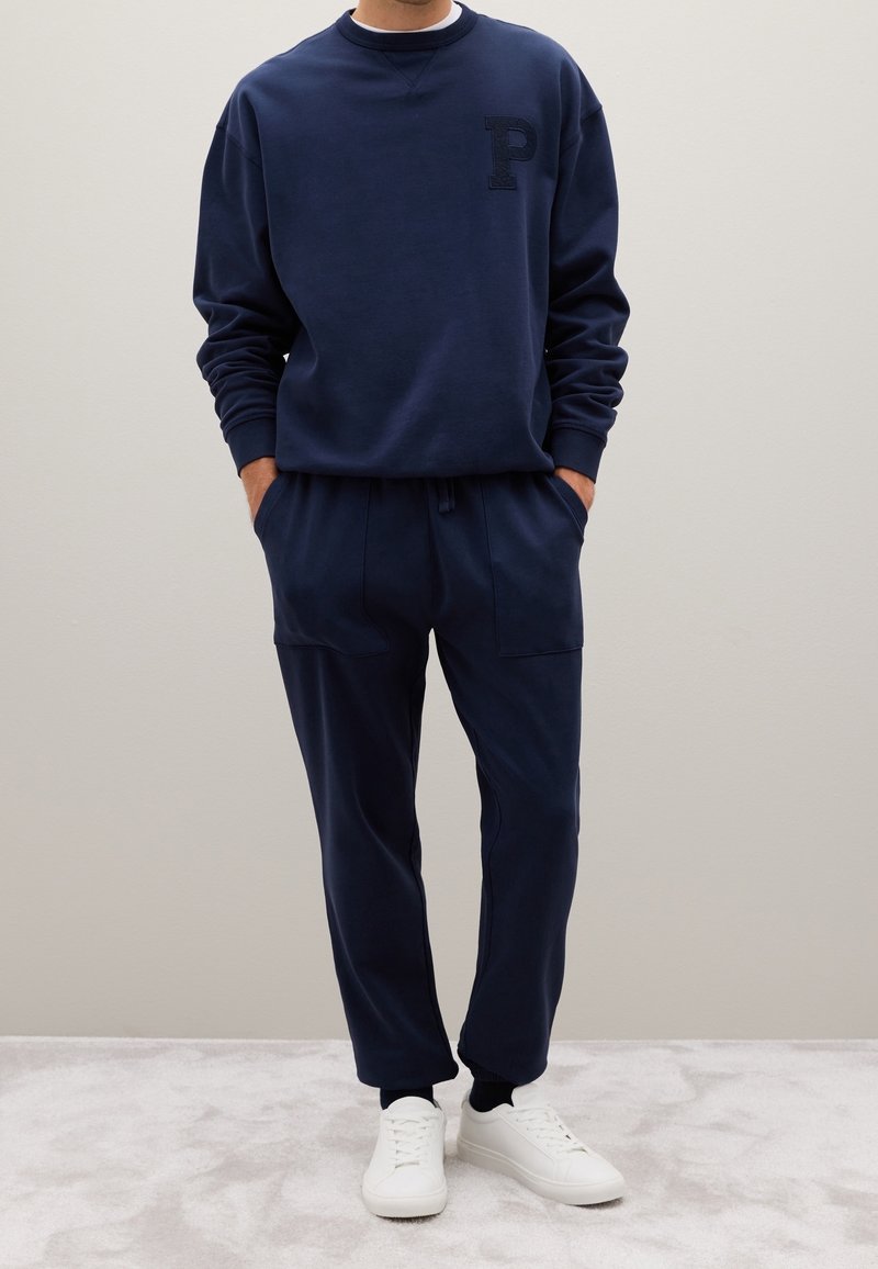 Homme portant un sweat-shirt bleu marine avec un « P » sur la poitrine, un pantalon de jogging assorti et des baskets blanches, debout les mains dans les poches.
