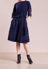 Mujer vestida con un vestido de encaje azul marino con patrones florales, mangas tres cuartos, un lazo en la cintura y botas negras hasta el tobillo, de pie sobre un suelo beige.
