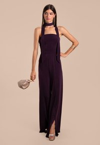 Mono morado sin mangas con un cuerpo ajustado, piernas anchas y fluidas, aberturas laterales y una corbata a juego en el cuello. La modelo sostiene un clutch ligero.