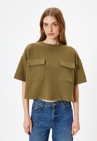 Olijfgroene cropped t-shirt met een ribbelstructuur, voorzien van twee klepzakken aan de voorkant en een ronde halslijn. Gecombineerd met blauwe jeans.