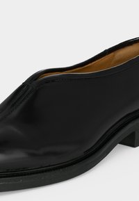 Chaussure de ville noire en cuir à enfiler avec un petit talon et une finition lisse, montrant les détails sur les côtés et le dessus sur fond uni.