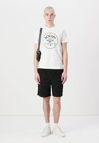 Bílý tričko s logem "JOOP! JEANS", v kombinaci s černými šortkami s potiskem a bílými teniskami. Model také nese malou černou kabelku přes rameno.
