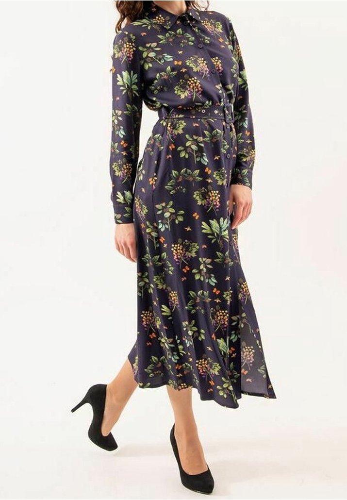 Femme portant une robe midi ceinturée à manches longues bleu marine à fleurs, associée à des chaussures à talons noirs, debout avec une jambe légèrement levée.