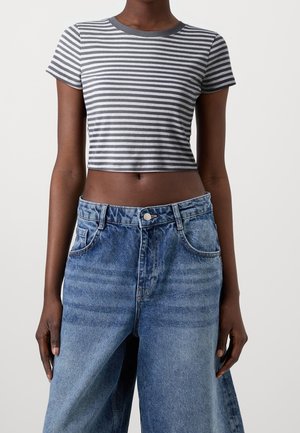 Person iført en grå og hvid stribet kort T-shirt og højtaljede løse blå jeans mod en ensfarvet baggrund.