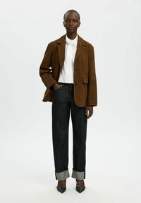 Blazer de ante marrón sobre una camisa blanca, combinada con vaqueros oscuros remangados en los puños. Tacones negros puntiagudos completan el conjunto.