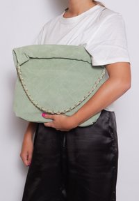 Groene suede handtas met een gequilt patroon, oversized vorm en een gouden kettinghandvat, vastgehouden tegen een witte achtergrond.