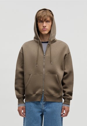 Junger Mann, der einen braunen Kapuzenpullover mit Reißverschluss über einem schwarz-weiß gestreiften Hemd und blauen Jeans trägt, vor einem schlichten Hintergrund stehend.