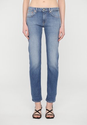 Kobieta w jasnoniebieskich dżinsach o prostych nogawkach i czarnych sandałach na wysokim obcasie z paskami, stojąca na tle jednolitego białego tła.