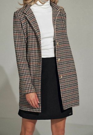 Vrouw draagt een beige, zwart en rood pied-de-poule jasje over een witte coltrui en een zwarte rok, staand tegen een neutrale achtergrond.