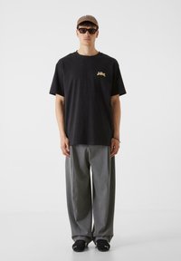 Melna kokvilnas T-kreklu ar zelta grafiku, kombinēts ar pelēkiem platām biksēm. Aksesuāri ietver brūnu cepuri un melnas sporta apavus.