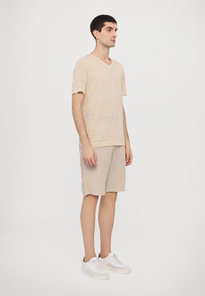 Beiges Leinen-T-Shirt mit V-Ausschnitt, kombiniert mit beigen Leinen-Shorts. Das Modell trägt weiße Sneaker und steht vor einem schlichten weißen Hintergrund.