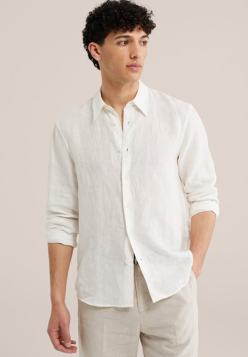 Homme portant une chemise blanche texturée à boutons avec les manches retroussées, pantalon beige, regardant sur le côté avec une main dans la poche.