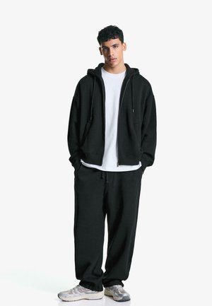 Črna bombažna zip-up kapuca s sprednjimi žepi, v kombinaciji s skladnimi črnimi trenirkami. Pod njo bela majica, sivi superge.