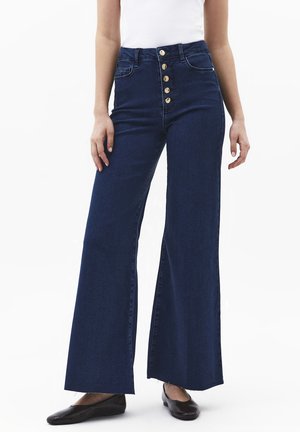 Vrouw die donkerblauwe wijdvallende jeans met hoge taille draagt, voorzien van vijf gouden knopen, en zwarte platte schoenen, staand tegen een witte achtergrond.