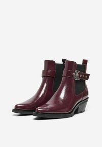 Un par de brillantes botines de cuero de color rojo oscuro con paneles elásticos laterales negros y una correa decorativa con hebilla sobre el tobillo, montados sobre tacones bajos en bloque.