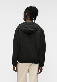 Μαύρο μπουφάν με κουκούλα και ελαστική ρίγα στη μέση και στα μανίκια, με ομαλή υφή και διακριτική ετικέτα της μάρκας στο αριστερό μανίκι.