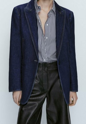 Personne portant un blazer en denim bleu foncé par-dessus une chemise à rayures verticales noires et blanches, et un pantalon en cuir noir taille haute.