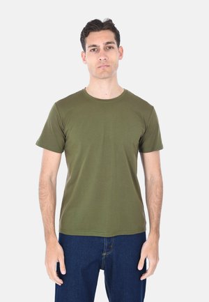 Giovane uomo in piedi, indossa una maglietta verde oliva a maniche corte e jeans blu scuro, rivolto in avanti su uno sfondo chiaro e uniforme.