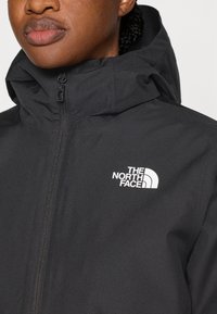 Μαύρο σακάκι με φερμουάρ και κουκούλα, κατασκευασμένο από ελαφρύ υλικό, με λευκό λογότυπο στο πάνω αριστερό μέρος του στήθους.