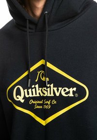Sudadera negra con un logo amarillo en forma de diamante que dice "Quiksilver" y "Original Surf Co. Desde 1969." Cuenta con cordones ajustables.