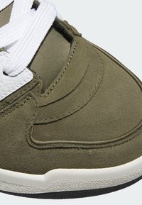 Green suede sneaker with white textured leather accents, featuring round white laces, smooth rubber sole, and detailed stitching.