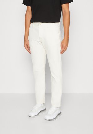 Une personne se tient debout, portant un t-shirt noir et un pantalon blanc, associé à des baskets blanches élégantes, sur un fond uni.