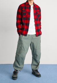Chemise en flanelle à carreaux rouge et noir, t-shirt blanc à col rond, pantalon cargo vert et baskets noires avec accents rouges. Tenue décontractée.