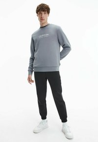 Felpa grigia con collo a giro e logo bianco "Calvin Klein" sul davanti, abbinata a pantaloni neri a sigaretta e sneaker bianche.