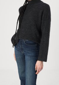 Personne portant un pull en maille gris foncé avec des poignets côtelés et un jean en denim bleu, debout devant un fond clair uni.
