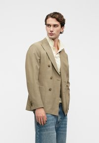 Άνδρας ντυμένος με μπεζ διπλόσπαστο σακάκι, κρεμ πουκάμισο με ανασηκωμένο γιακά και μπλε τζιν, κοιτάει αριστερά σε έναν απλό φόντο.