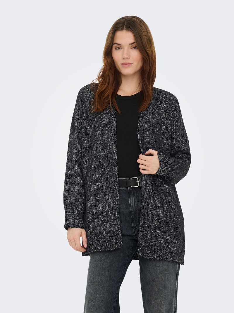 Woman with brown hair wearing a black shirt, dark speckled open cardigan, and black belt with dark gray jeans, standing against a plain background.