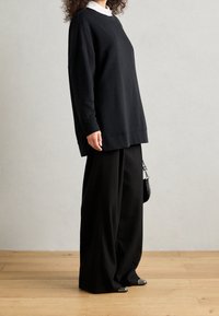 Czarna, oversize bluza z długimi rękawami, zestawiona z szerokimi czarnymi spodniami, na tle jasnej drewnianej podłogi i neutralnego tła.