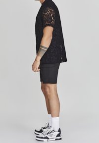 Camicetta nera in pizzo a maniche corte con una vestibilità rilassata, abbinata a pantaloni corti neri. Indossata con calzini bianchi e sneakers nere e bianche.