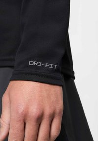 Musta pitkähihaurheilupaita, jossa hopeanvärinen DRI-FIT-logo hihansuutessa. Sileä, joustava kangas, istuva muoto, minimaliset ompelut.