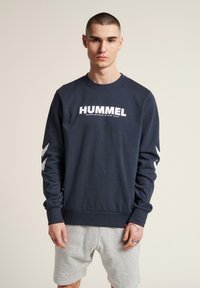 Felpa blu navy con logo bianco "HUMMEL", colletto e polsini a coste, con dettagli a chevron bianchi sulle maniche. Indossata con pantaloni corti grigi.