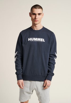 Navyblauwe sweatshirt met witte "HUMMEL" logo, geribbelde halslijn en boorden, met witte chevronaccenten op de mouwen. Draagt met grijze shorts.